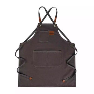 Könnte beinhalten: Eine dunkelgraue Schürze aus Canvas mit verstellbaren Trägern und zwei Taschen. Die Schürze hat braune Lederakzente und ist zum Kochen oder Servieren gedacht.