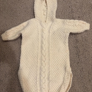 May include: A cream-colored, long-sleeved hooded garment featuring a central vertical cable knit pattern. The piece is crafted with a textured knit stitch throughout the body and sleeves, finished with ribbed cuffs and a rounded bottom hemline.