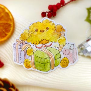 Puede incluir: Una pegatina de acuarela con un perro amarillo y esponjoso que sostiene una caja de regalo verde con una cinta naranja. El perro está rodeado de otros regalos en rosa y morado. La pegatina tiene un tema festivo y navideño.