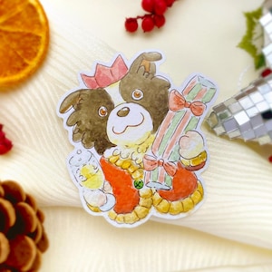 Op de afbeelding: Een aquarelsticker van een hond met een rode kroon die een ingepakt cadeau vasthoudt. De hond heeft bruine vacht en draagt een rode en gele outfit. Het cadeau is gestreept met een rode strik. Een klein glas vloeistof zit in de andere poot van de hond.