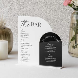 May include: A wedding bar sign with a white and black design. The sign includes a list of drinks and a section for signature drinks. A vase of flowers and candles are in the background.
