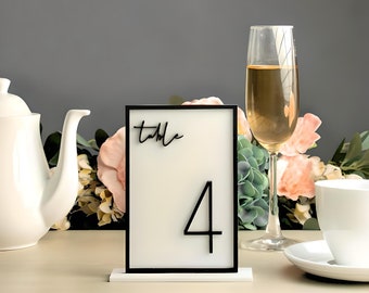 May include: Black and white table number sign with the text "table" and the number "4" on a white background. The sign is on a white stand.
