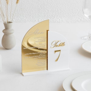Peut inclure: Un numéro de table et un menu en acrylique or et blanc. Le menu a un dessus doré en forme d'arche avec les plats listés. Le numéro de table est blanc avec un design "Table 7" doré. Le menu comprend des entrées, des plats et des plats principaux.