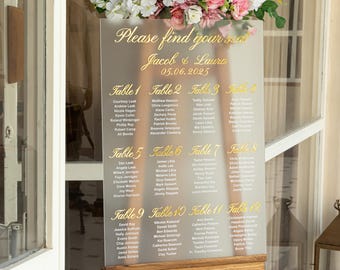 Könnte beinhalten: Ein transparenter Acryl-Sitzplan für eine Hochzeit mit goldenen Buchstaben. Der Plan listet die Namen der Gäste auf, die den einzelnen Tischen zugeordnet sind. Der Plan ist mit rosa und weißen Blumen verziert.