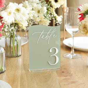 May include: Sage green table number 3 with white script text "Table". The acrylic sign is displayed on a wooden table with floral arrangements and glassware. Perfect for weddings and events.