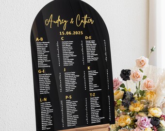 May include: Black acrylic arch-shaped wedding seating chart with gold lettering. The chart lists guest names alphabetically by table number. The chart is on a wooden easel.