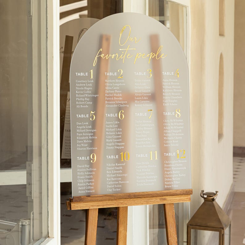 Puede incluir: Un plano de asientos de boda de acr&iacute;lico transparente con letras doradas y un caballete de madera. El texto "Our favorite people" est&aacute; en la parte superior. El gr&aacute;fico enumera los nombres bajo los n&uacute;meros de mesa del 1 al 12.