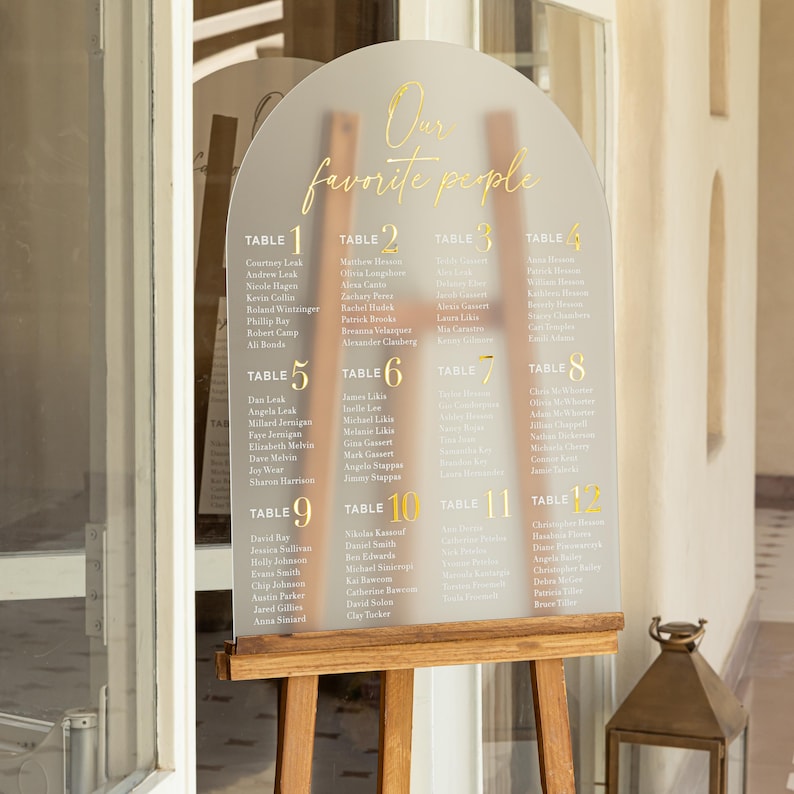 Puede incluir: Un plano de asientos de boda de acr&iacute;lico transparente con letras doradas y un caballete de madera. El texto "Our favorite people" est&aacute; en la parte superior. La tabla enumera nombres bajo los n&uacute;meros de mesa del 1 al 12.