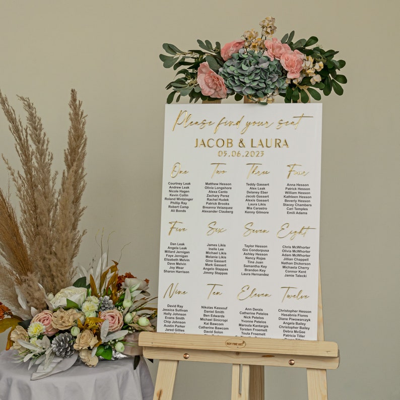 K&ouml;nnte beinhalten: Eine wei&szlig;e Hochzeitsplatzkarte mit goldenen Buchstaben. Die Karte tr&auml;gt den Titel "Bitte finden Sie Ihren Platz" und listet die Namen der G&auml;ste f&uuml;r jede Tischnummer auf. Die Karte wird auf einem Holzst&auml;nder mit einem Blumenarrangement davor angezeigt.