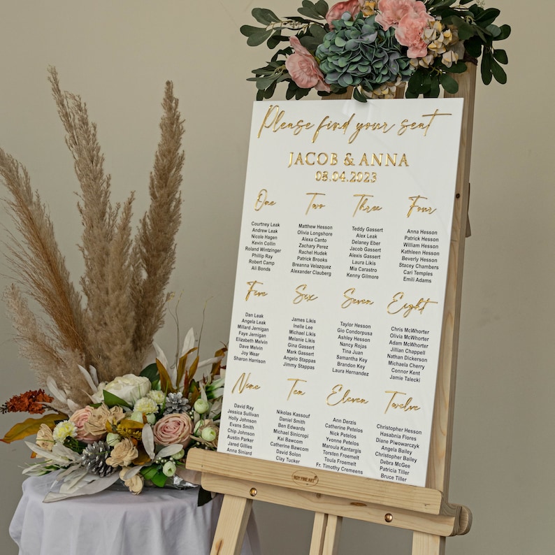 K&ouml;nnte beinhalten: Ein wei&szlig;er Sitzplan mit goldenen Buchstaben f&uuml;r einen Hochzeitsempfang. Der Plan tr&auml;gt den Titel "Bitte finden Sie Ihren Platz" und listet die Namen der G&auml;ste f&uuml;r jeden Tisch auf, die von eins bis zw&ouml;lf nummeriert sind. Der Plan wird auf einem Holzst&auml;nder angezeigt.