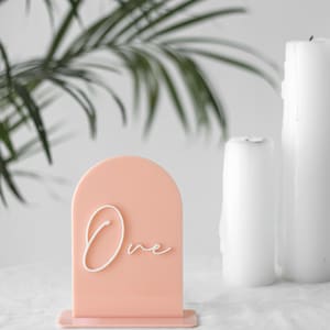 Puede incluir: Un n&uacute;mero de mesa de acr&iacute;lico de color melocot&oacute;n con una inscripci&oacute;n cursiva blanca que dice "Uno". El n&uacute;mero de mesa est&aacute; sobre una superficie blanca con dos velas blancas en el fondo.
