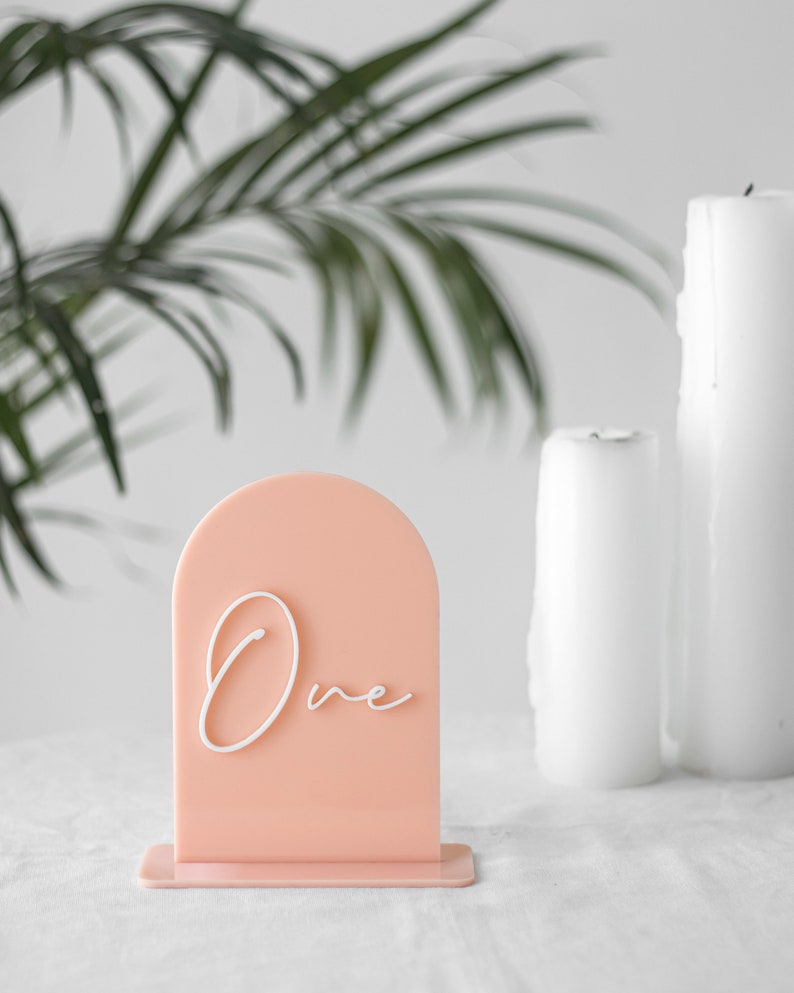Puede incluir: Un n&uacute;mero de mesa de acr&iacute;lico de color melocot&oacute;n con una inscripci&oacute;n cursiva blanca que dice "Uno". El n&uacute;mero de mesa est&aacute; sobre una superficie blanca con dos velas blancas en el fondo.