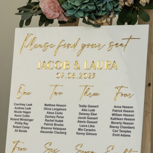K&ouml;nnte beinhalten: Wei&szlig;e Hochzeitssitzordnung mit goldenen Buchstaben. Die Karte listet die Namen der G&auml;ste f&uuml;r jeden Tisch auf, nummeriert von eins bis vier. Oben auf der Karte steht "Bitte finden Sie Ihren Platz" und "Jacob & Laura 05.06.2023".