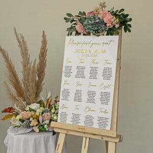 K&ouml;nnte beinhalten: Wei&szlig;e Hochzeitssitzordnung mit goldener Beschriftung auf einem Staffelei. Die Karte listet die Namen der G&auml;ste nach Tischnummer auf. Der Text lautet "Bitte finden Sie Ihren Platz Jacob & Laura 07.06.2023".