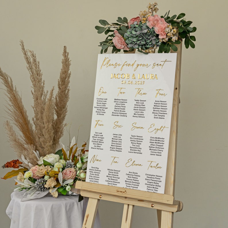 K&ouml;nnte beinhalten: Wei&szlig;e Hochzeitssitzordnung mit goldener Beschriftung auf einem Staffelei. Die Karte listet die Namen der G&auml;ste nach Tischnummer auf. Der Text lautet "Bitte finden Sie Ihren Platz Jacob & Laura 07.06.2023".