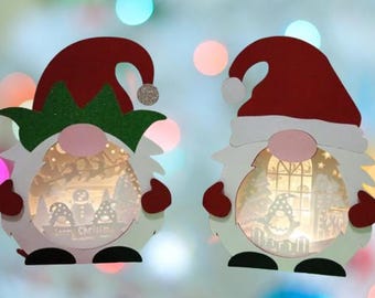 Pode incluir: Dois gnomos de Natal com janelas iluminadas. O gnomo à esquerda tem um chapéu verde e o gnomo à direita tem um chapéu vermelho. Ambos os gnomos têm barba branca e estão segurando um coração vermelho.