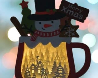 May include: A wooden Christmas ornament shaped like a mug with a snowman inside. The mug has a red rim and handle. The snowman is wearing a black top hat and a red and white scarf. The ornament has a light inside that illuminates a winter scene with trees and houses.