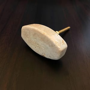 May include: An oval-shaped cabinet knob made of a light beige, porous stone material. The knob features a textured, natural surface with subtle colour variations and is attached to a threaded brass-coloured metal bolt with a small hexagonal nut.