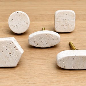 May include: A collection of five cabinet knobs made from off-white, porous stone with natural pitting. The set includes one circular knob, one square knob, one hexagonal knob, and two elongated oval knobs, each featuring a brass-toned metal screw.