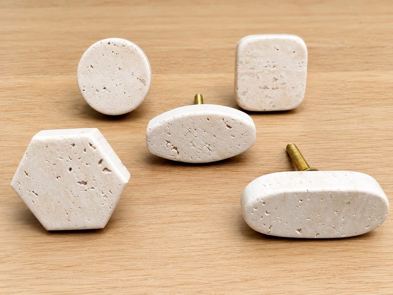 May include: A collection of five cabinet knobs made from off-white, porous stone with natural pitting. The set includes one circular knob, one square knob, one hexagonal knob, and two elongated oval knobs, each featuring a brass-toned metal screw.
