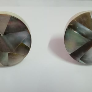 May include: Two round decorative cabinet knobs. Each knob is composed of a mosaic of iridescent, geometric shapes in shades of gray, brown, and green. The knobs are set against a white background.