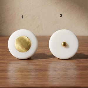 Könnte beinhalten: Zwei runde weiße Knöpfe mit der Beschriftung 1 und 2. Knopf 1 hat eine große, gebürstete goldene kreisförmige Einlage in der Mitte. Knopf 2 hat einen kleinen, polierten goldenen kugelförmigen Knopf in der Mitte. Beide Knöpfe sind mattweiß.