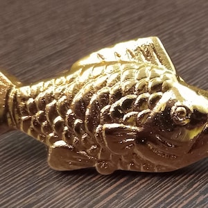 May include: A gold-colored fish figurine with detailed scales, fins, and tail. The fish is positioned horizontally, with its head to the right. The figurine is made of a shiny, metallic material and is set against a dark, wooden surface.