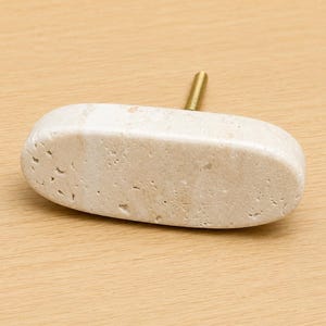 May include: An oval-shaped cabinet knob made of light beige natural stone with a porous, textured surface. The knob features a single gold-toned metal threaded bolt extending from the back, designed for attachment to furniture or cabinetry doors.