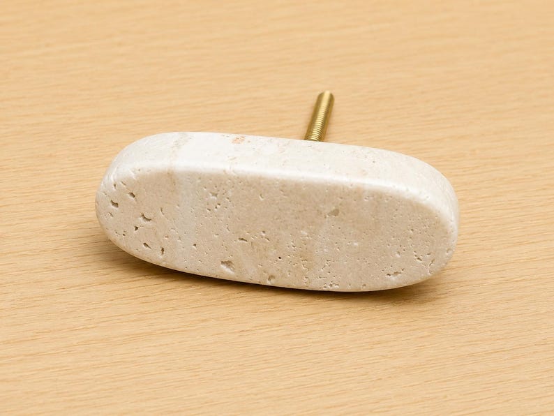 May include: An oval-shaped cabinet knob made of light beige natural stone with a porous, textured surface. The knob features a single gold-toned metal threaded bolt extending from the back, designed for attachment to furniture or cabinetry doors.