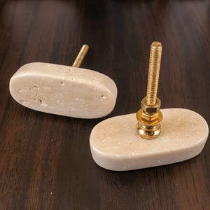 May include: Two oval-shaped cabinet knobs made of smooth, light beige stone with natural surface pitting. Each knob features a central gold-toned metal bolt with a threaded shaft, a flat washer, and a hexagonal nut for secure installation.