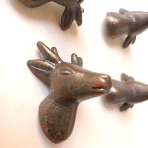 May include: Close-up of several bronze-colored deer head cabinet knobs. The knobs are detailed with antlers, eyes, and a textured surface. The deer heads are arranged against a white background, creating a rustic aesthetic.