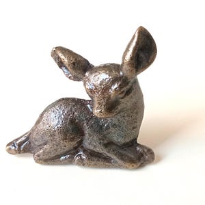 May include: A bronze-colored figurine of a resting deer. The deer is in a seated position with its head lowered and ears perked up. The figurine has a textured surface and is set against a white background.