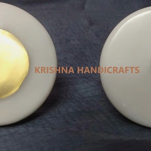 Könnte beinhalten: Zwei weiße runde Knöpfe mit goldenen Akzenten. Der erste Knopf hat einen großen goldenen Kreis in der Mitte. Der zweite Knopf hat eine kleine goldene Kugel in der Mitte.  KRISHNA HANDICRAFTS ist auf dem Bild gedruckt.