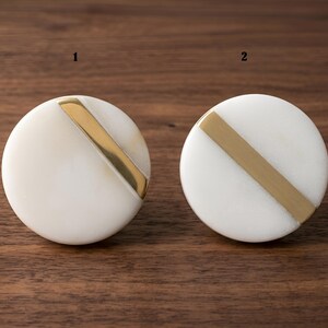Peut inclure: Deux boutons circulaires étiquetés 1 et 2, chacun présentant une surface en pierre blanche lisse coupée par une bande diagonale en métal doré poli. Les boutons sont ronds avec une face plate et un design géométrique net sur la surface avant.