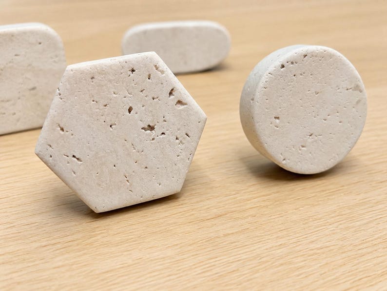 May include: A collection of four cabinet knobs made from off-white porous stone. The set includes one hexagonal knob, one circular knob, and two elongated oval knobs, all featuring a natural, textured surface with small indentations.