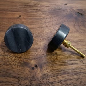 May include: Two circular cabinet knobs featuring a dark gray and black marbled stone surface. Each knob is attached to a gold-toned metal bolt with a matching nut, designed for mounting onto furniture or cabinetry doors and drawers.