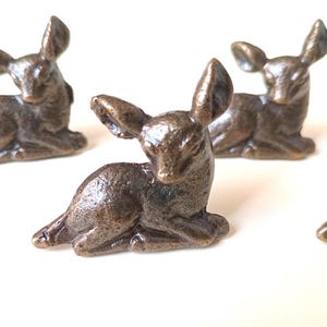 Puede incluir: Figuras de ciervos de color bronce en una pose relajada y sentada. Las pequeñas esculturas tienen detalles, incluyendo orejas grandes y una superficie texturizada. Las figuras están dispuestas sobre una superficie blanca, algunas enfocadas y otras borrosas.
