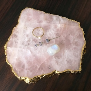 May include: A pale pink rose quartz slab with a jagged, gold-painted edge. Resting on the surface is a thin gold ring with a small white pearl, a single sprig of dried lavender, and a smooth, translucent white moonstone with a blue sheen.