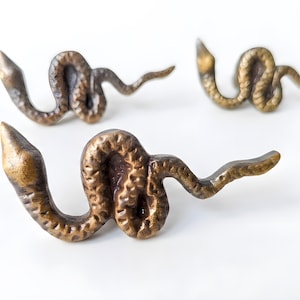 May include: Three brass-toned metal drawer knobs shaped like coiled snakes. Each knob features a textured, scaled surface with a pointed head and a winding body. The finish is a muted, antique bronze color with darker recessed areas.