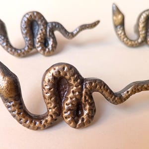 May include: Close-up of several bronze-colored snake-shaped cabinet pulls. The pulls are detailed with textured scales and curved bodies, suggesting a decorative hardware design. The snake heads are pointed, and the tails are slightly curved.
