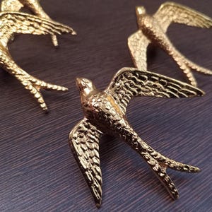 May include: Close-up of several gold-colored swallow figurines with outstretched wings. The birds are detailed with textured feathers and are arranged on a dark, possibly wooden, surface. The lighting highlights the metallic sheen.