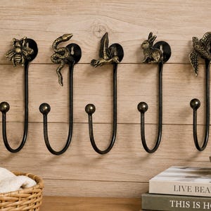 Puede incluir: Cinco ganchos de pared de metal con acabado en bronce oscuro. Cada gancho presenta una figura animal única en la parte superior, incluyendo una abeja, una serpiente, un pájaro en vuelo, un conejo y una golondrina. Diseño largo y curvado.