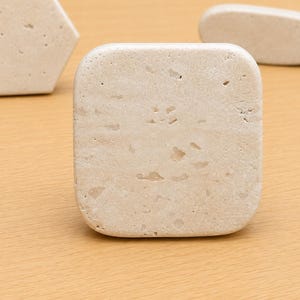 May include: A square-shaped beige stone tile with a porous, textured surface featuring natural indentations and variations in color. The tile has rounded corners and a smooth, matte finish, showcasing the organic, earthy appearance of the natural stone material.