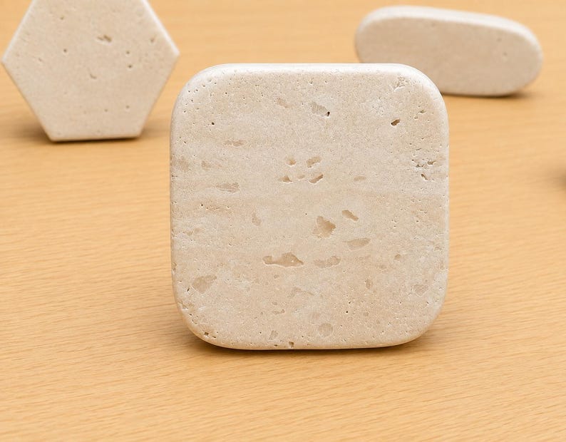 May include: A square-shaped beige stone tile with a porous, textured surface featuring natural indentations and variations in color. The tile has rounded corners and a smooth, matte finish, showcasing the organic, earthy appearance of the natural stone material.