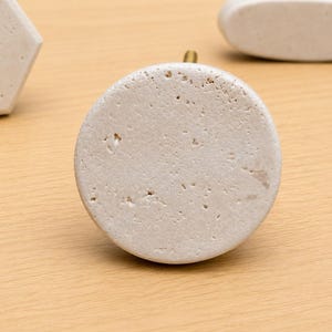 May include: A circular cabinet knob made of off-white, porous stone with a textured, pitted surface. The knob features a small, metallic gold-colored post extending from the back, centered behind the round stone face.