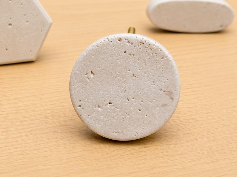 May include: A circular cabinet knob made of off-white, porous stone with a textured, pitted surface. The knob features a small, metallic gold-colored post extending from the back, centered behind the round stone face.