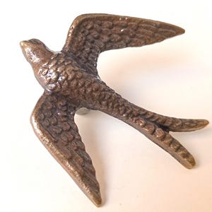 Może przedstawiać: Brązowa figurka ptaka w locie. Ptak ma rozłożone skrzydła i szczegółową, teksturowaną powierzchnię. Figurka jest prawdopodobnie wykonana z metalu i ma być ozdobnym elementem.