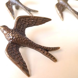 Könnte beinhalten: Dekorative Hardware in Form von bronzefarbenen Schwalben. Die Vögel sind im Flug mit ausgebreiteten Flügeln und detaillierten Federn dargestellt. Die Hardware ist wahrscheinlich für einen Schrank oder eine Schublade.