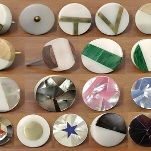 May include: A collection of twenty circular cabinet knobs featuring diverse designs including inlaid brass, green malachite, iridescent mother-of-pearl, wood, and geometric patterns. Each knob has a smooth, rounded face with varied textures and colors.