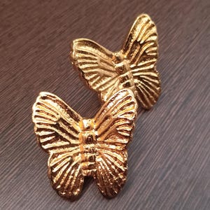 May include: Two gold-colored butterfly-shaped decorative objects. The butterflies have detailed wing patterns and are positioned on a dark, textured surface. The objects appear to be made of metal and are highly reflective.